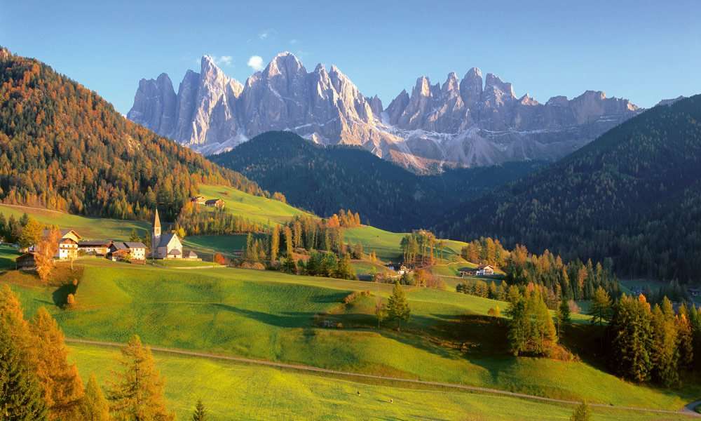 An alpine village with a white church spire on green meadows, beneath jagged pale dolomite peaks.