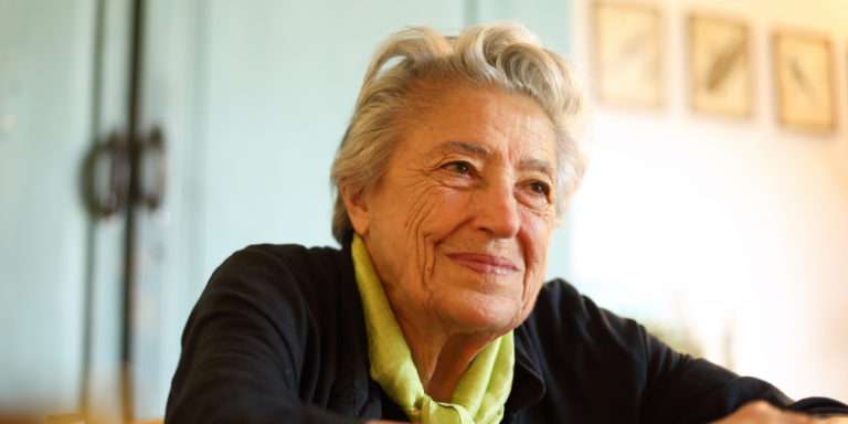 An older woman with short white hair seated indoors, turning toward the camera with a slight smile.