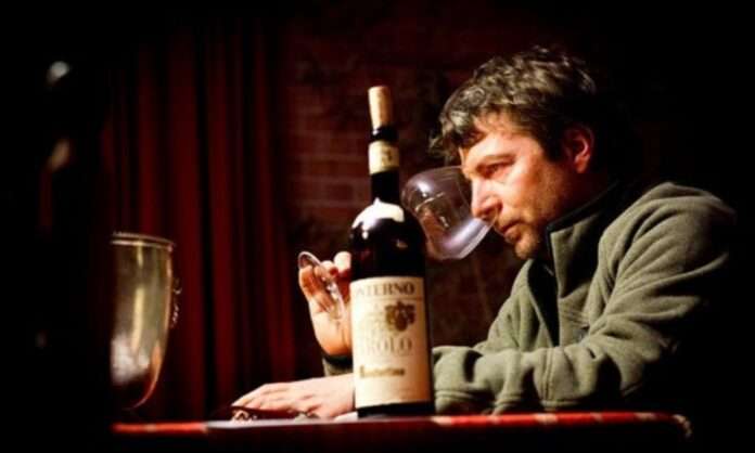 A man in a dark room leaning over a table with an old wine bottle and glass, lit by a single warm light.