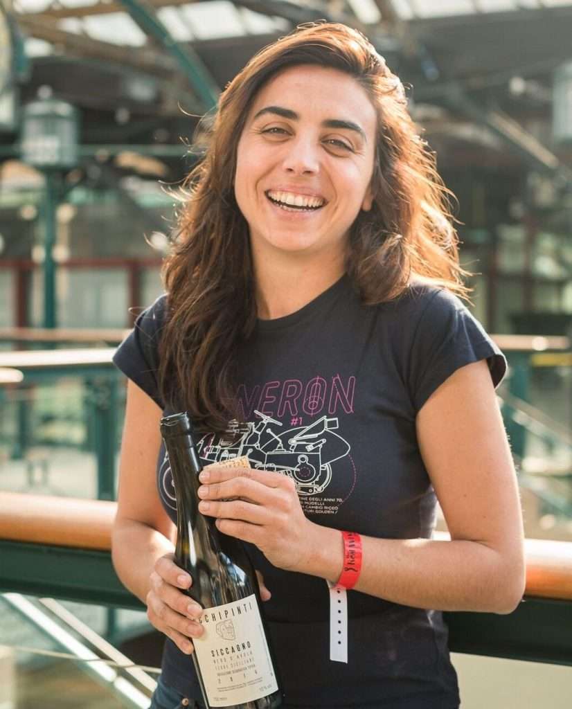 A woman with long hair in a dark T-shirt smiling and holding a bottle of wine inside a glass-roofed building.
