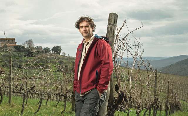 A man with curly hair in a red jacket standing among bare winter vines on a hilltop.