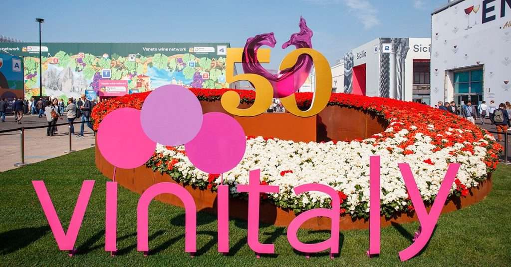 An outdoor trade-fair entrance with a large pink Vinitaly sign and flower bed, pavilions behind.