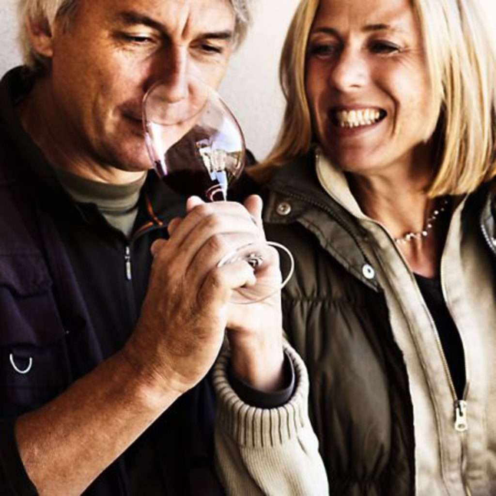 A man tasting from a wine glass beside a smiling blonde woman, both in winter jackets, photographed close up.