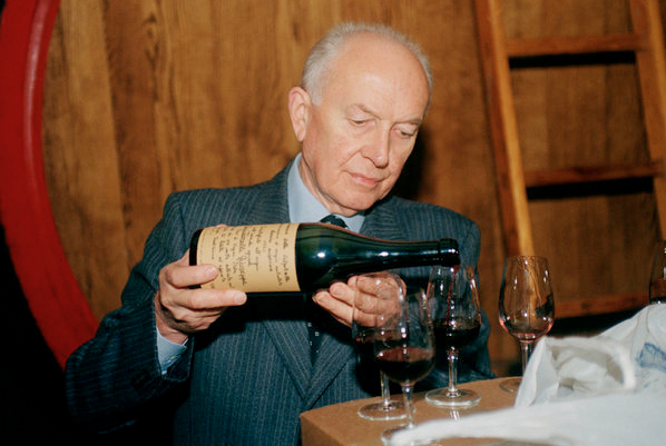 An elderly man in a suit pouring wine from an old bottle into glasses, standing beside a large wooden cask.