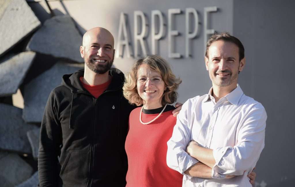 Three people, two men and a woman, standing together in front of a wall bearing the Ar.Pe.Pe. name.