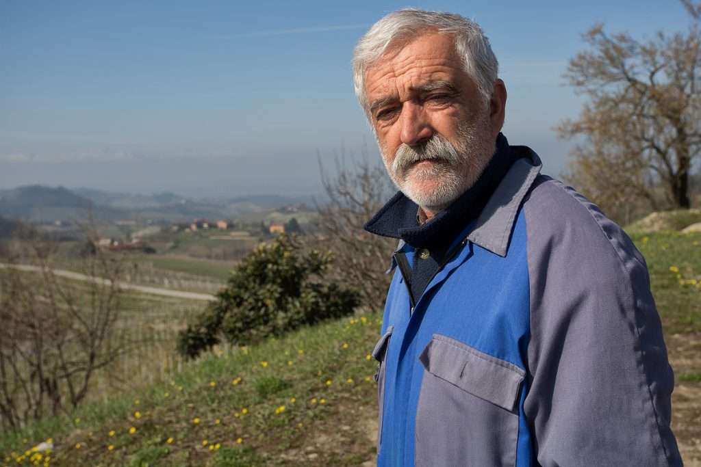 An older man with a white beard in a blue work jacket standing on a grassy slope above open countryside.