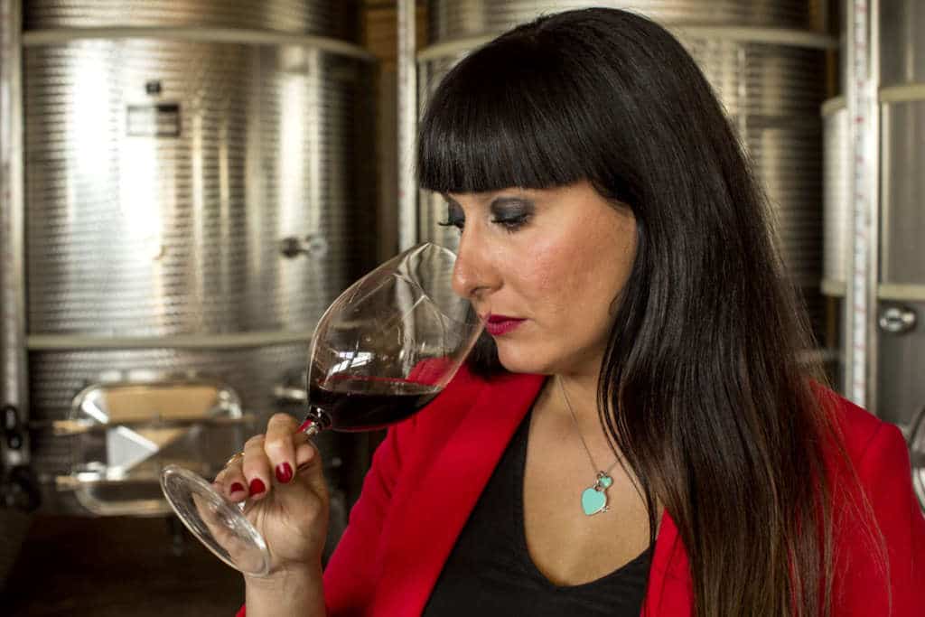 A woman in a red jacket tasting a glass of red wine in front of stainless steel tanks.