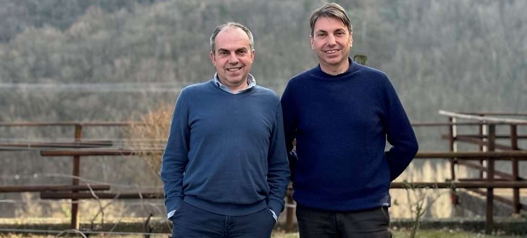 Two men standing side by side at a wooden fence with a wooded valley behind them.
