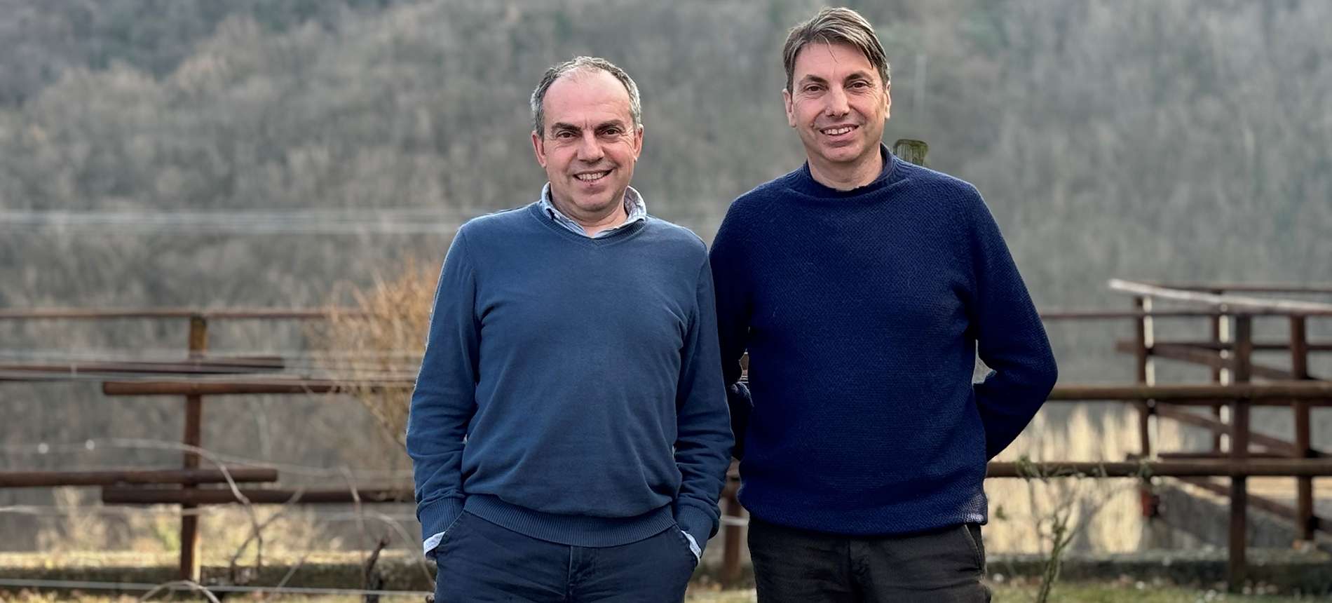 Two men standing side by side at a wooden fence with a wooded valley behind them.