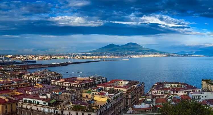A bay city seen from above, rooftops in the foreground and a broad volcano rising across the water.