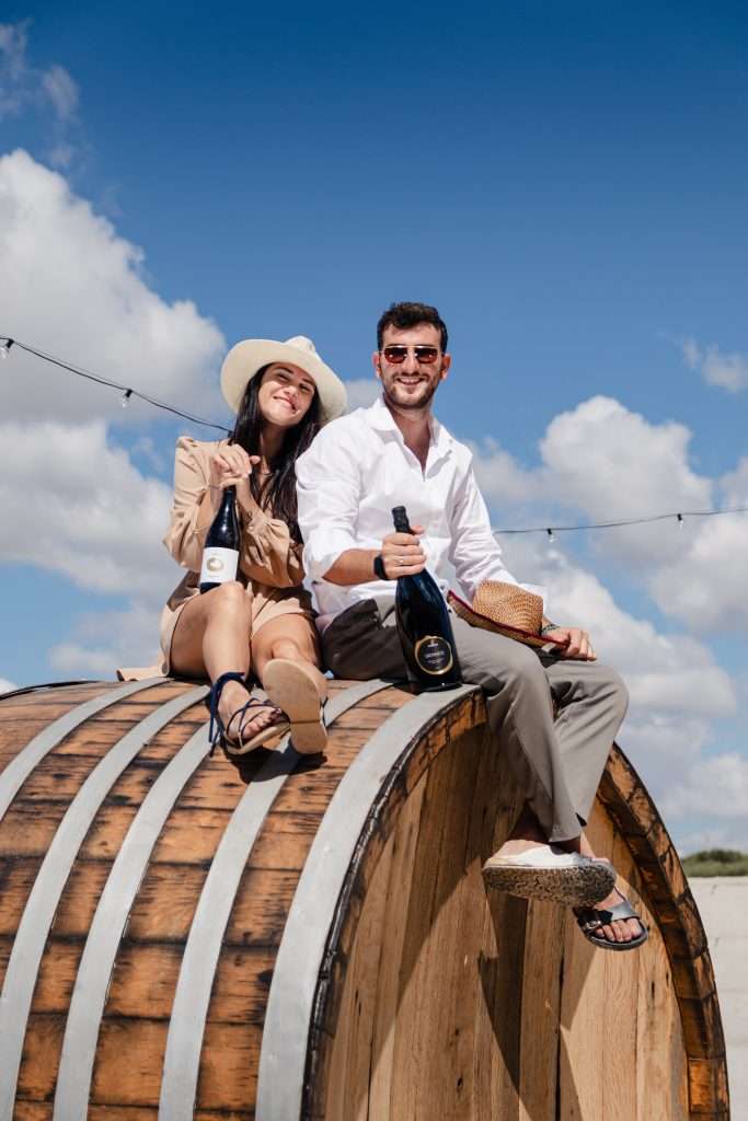 A young couple sitting on top of a very large wooden cask outdoors under a bright blue sky, each holding a bottle.