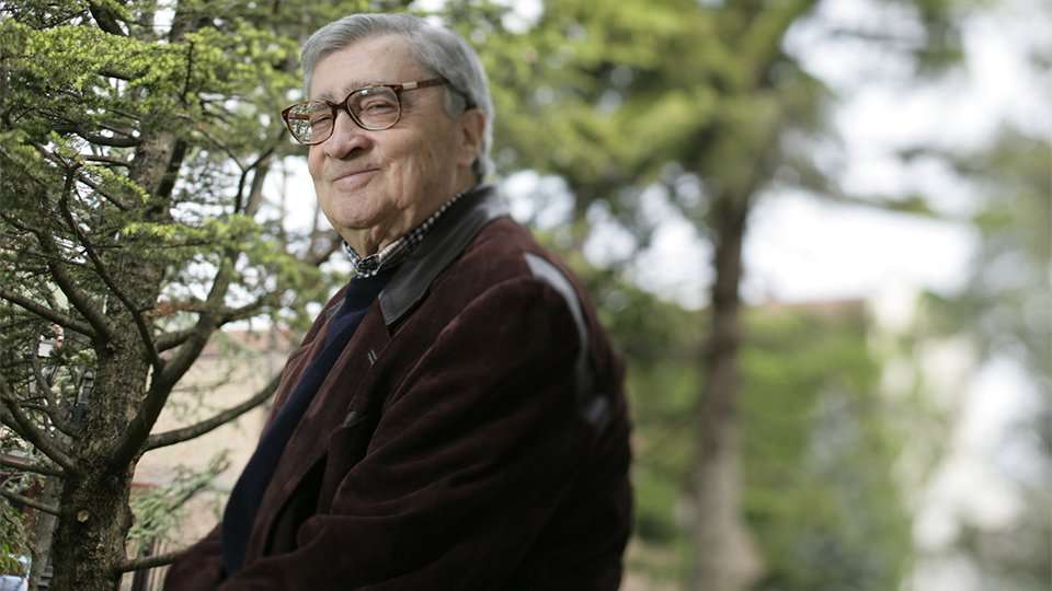 An elderly man in a tweed jacket and glasses seated outdoors among pine trees, looking into the distance.