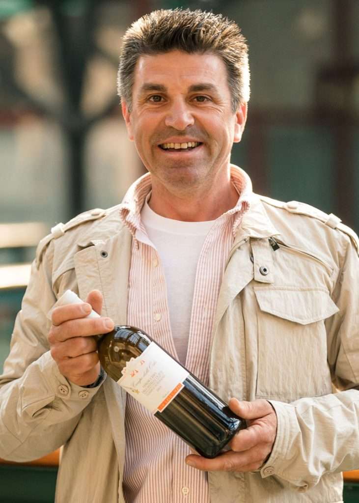 A smiling man in a pale jacket holding a bottle of wine horizontally toward the camera.