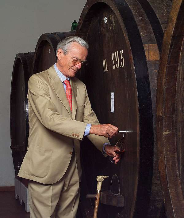 An elderly man in a pale suit drawing wine from the tap of a very large wooden cask.