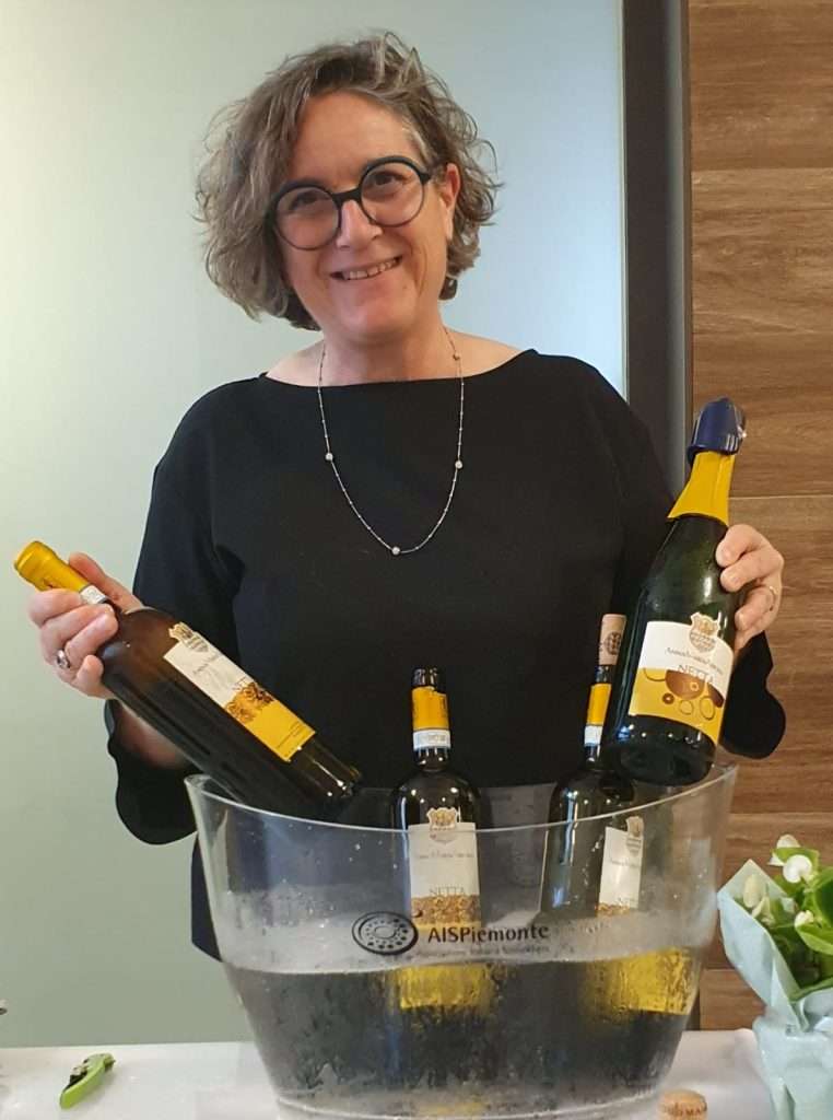 A woman with short curly hair and glasses holding two bottles of white wine above an ice bucket of bottles.