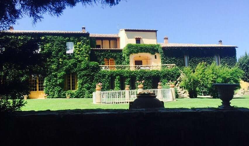 A long ivy-covered villa with a tiled roof behind a clipped lawn and stone urns.