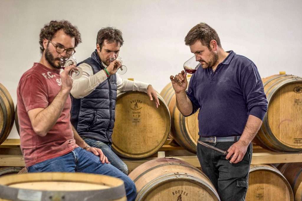 Three men tasting wine from glasses while leaning on stacked barrels in a cellar.