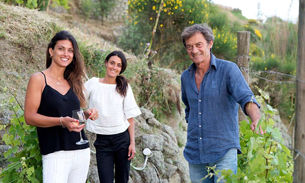 Two women holding wine glasses and a man in a blue shirt standing together on a steep terraced vineyard.