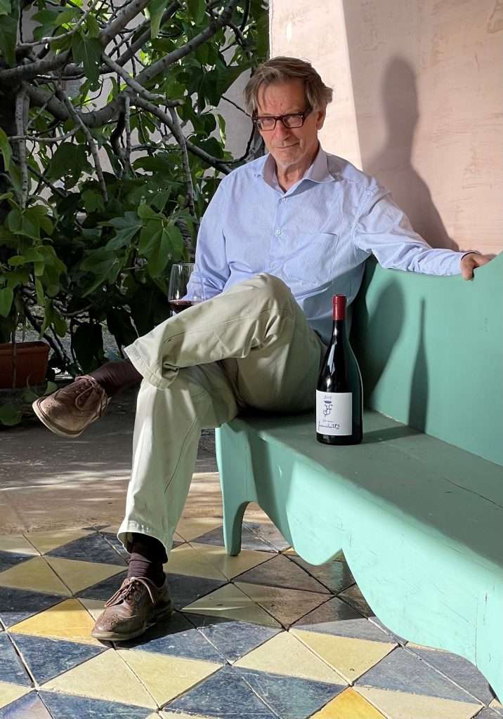 A man in a pale shirt sitting on a green bench on a tiled terrace, a bottle of wine beside him.