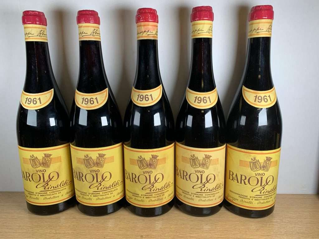 Five bottles of 1961 Barolo with yellowed labels standing in a row against a pale wall.