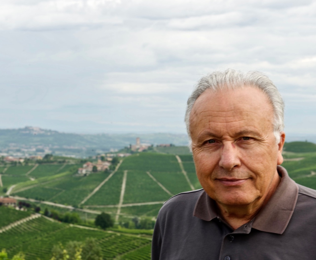 A man with grey hair in a dark polo shirt, vineyard hills stretching away behind him.