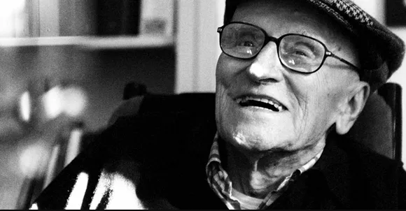 A black and white photograph of an elderly man in a flat cap and glasses, smiling.