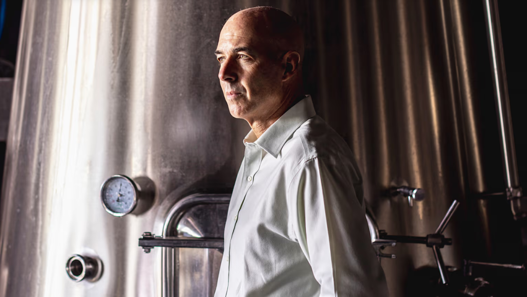 A man in a white shirt standing between stainless steel tanks in a cellar.