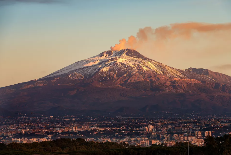 A volcano at sunrise with a plume of smoke from the summit, a lit city spread below.