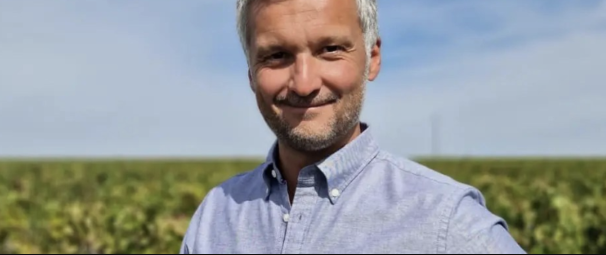 A man in a blue shirt smiling in front of a vineyard under a pale sky.
