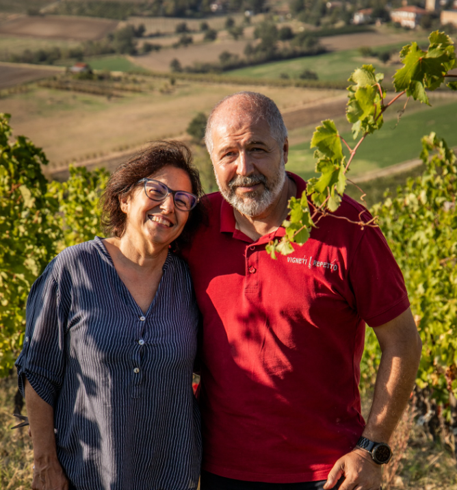 A woman and a man in a red polo shirt standing arm in arm at the edge of a vineyard.