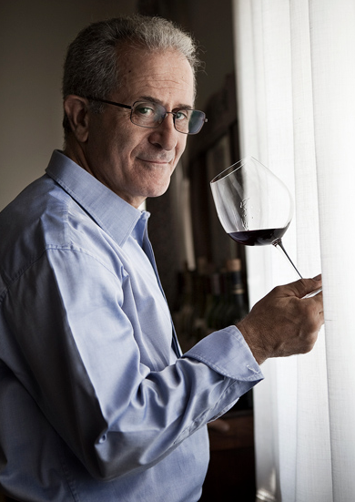 A man in a pale shirt and glasses holding a glass of red wine up to the light.