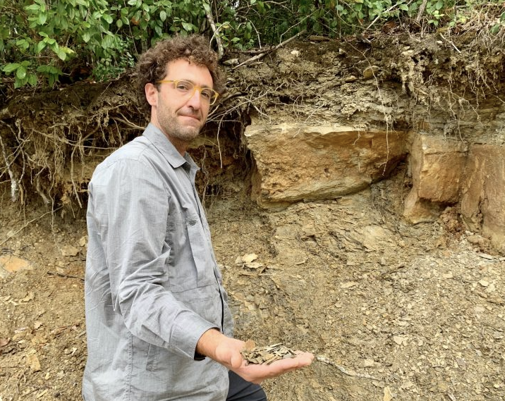 A man crouched beside a road cutting holding a handful of soil, exposed rock layers behind him.