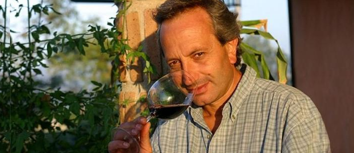 A man tasting a glass of red wine outdoors, foliage and a building behind him.