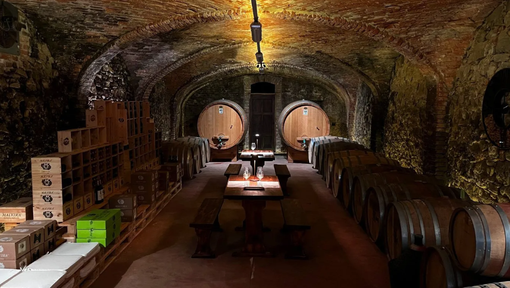 A vaulted stone cellar with large oak casks along one wall and cases stacked on the other.