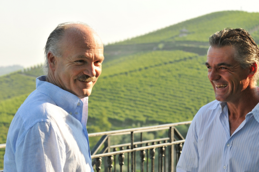 Two men leaning on a railing above a vineyard valley, both smiling at the camera.