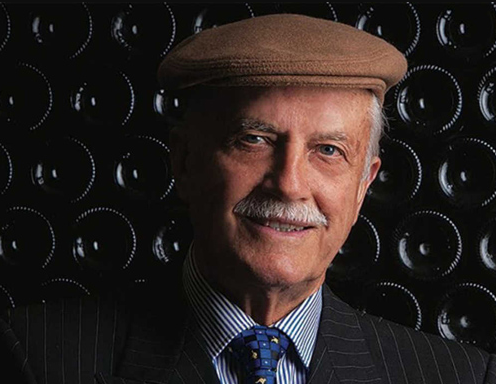 A man in a flat cap and pinstriped suit in front of a wall of stacked bottle bases.